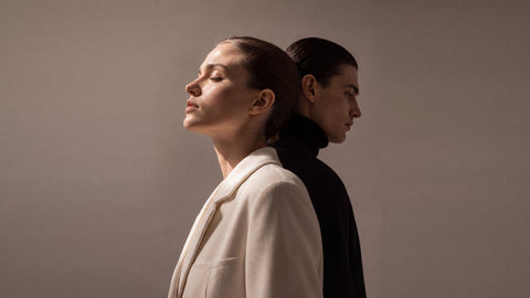 Woman in a cream coat and man in a black turtleneck standing back to back in profile, Grace’ Routine