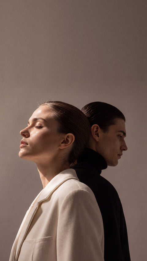 Woman in a cream coat and man in a black turtleneck standing back to back in profile, Grace’ Routine